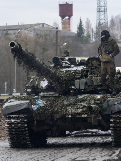Auf einem Panzer steht ein ukrainischer Soldat, gefolgt von weiteren Panzern. Im Hintergrund ist die ukrainische blau-gelbe Flagge zu sehen.