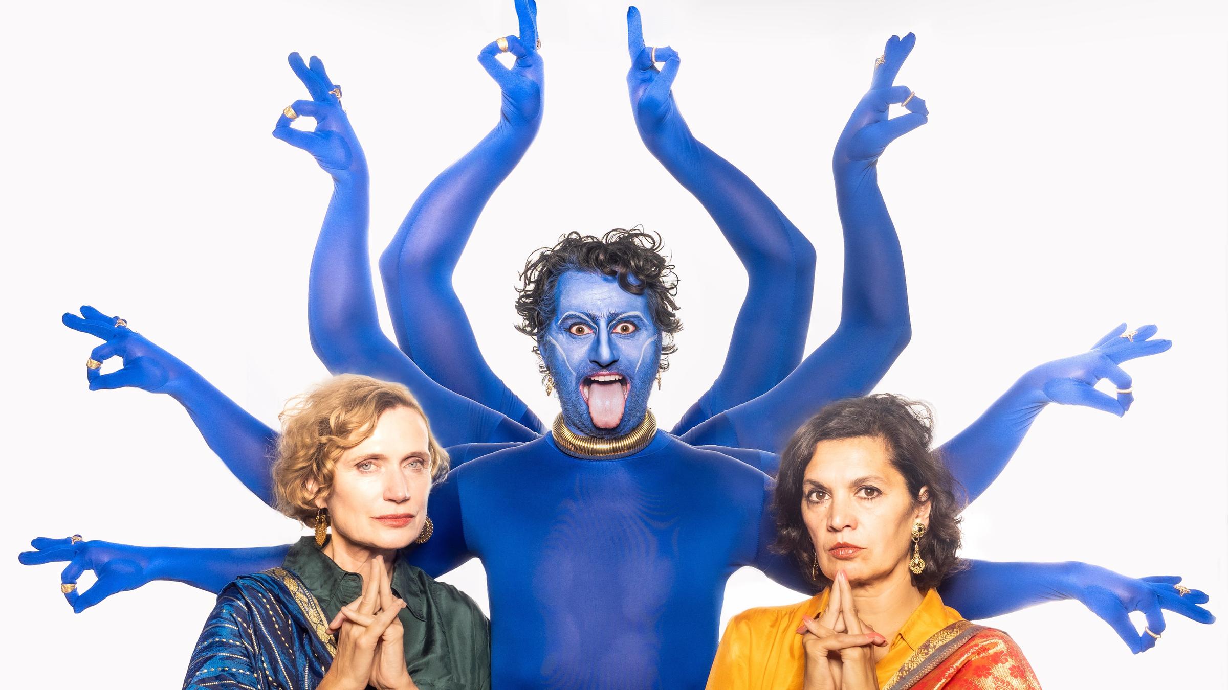 Ein Mann in Blau als indische Göttin mit acht Armen. Links und rechts daneben jeweils eine Frau in indisch anmutender Kleidung.