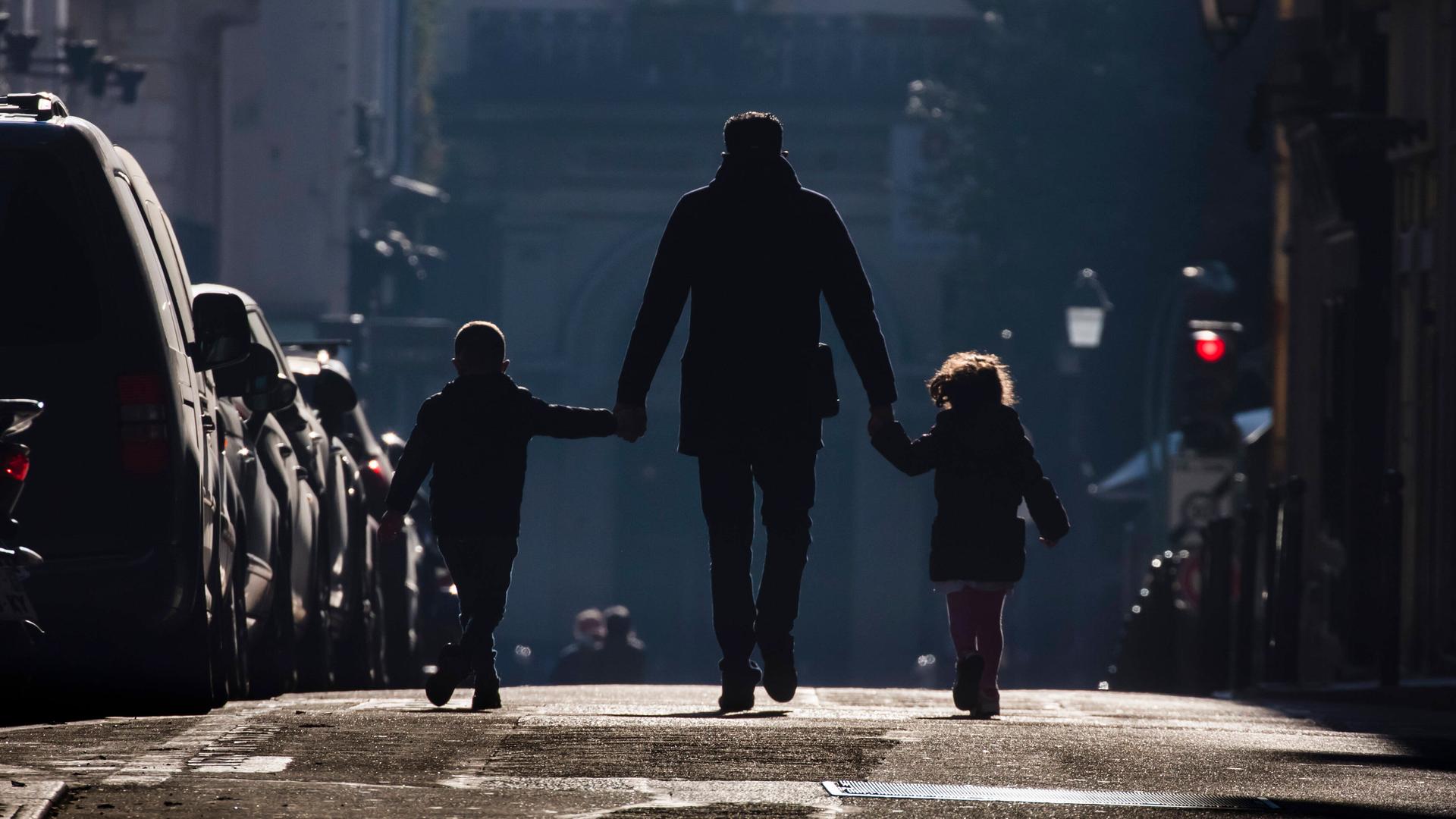 Ein Vater hält seine beiden Kinder an der Hand und geht mit beiden eine Straße entlang. Ein Vater hält seine beiden Kinder an der Hand und geht mit beiden eine Straße entlang.