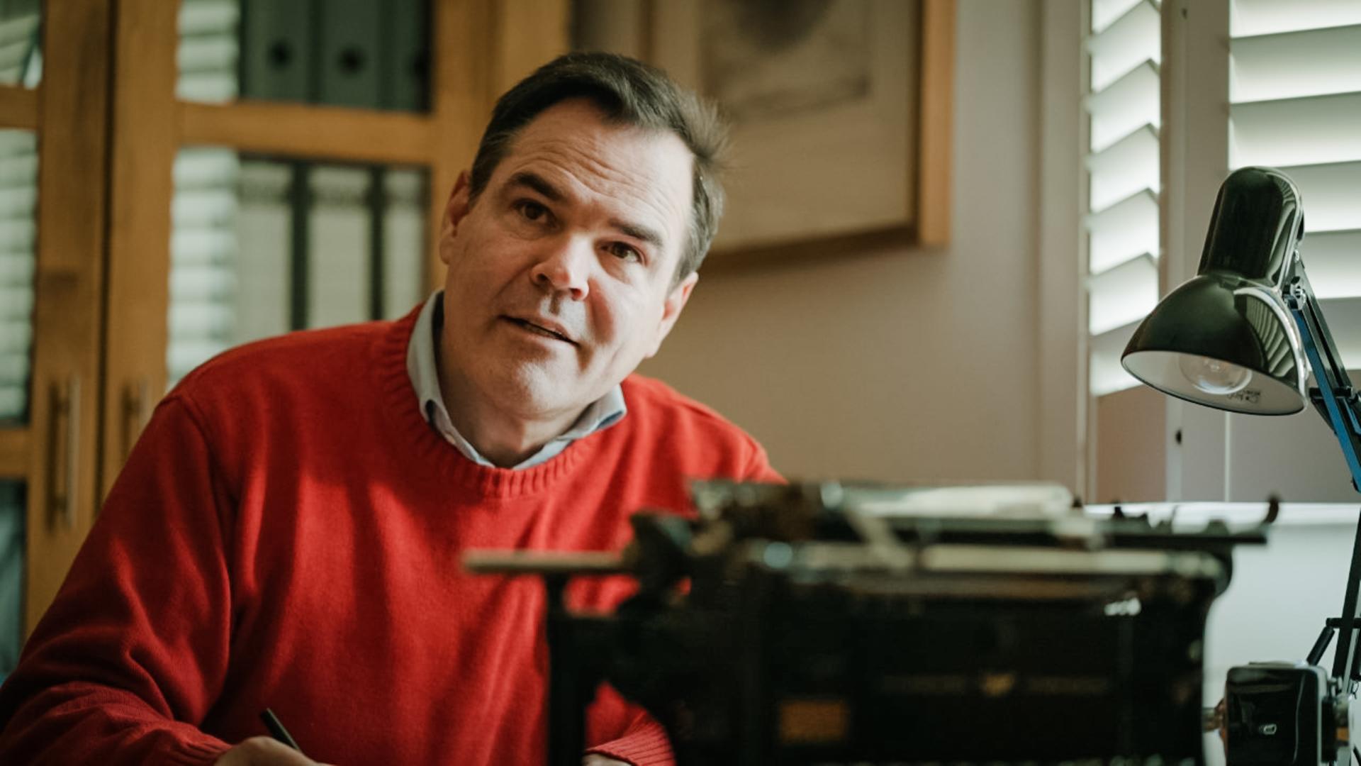 Uwe Tellkamp sitzt im roten Pullover vor einer mechanischen Schreibmaschine und scheint aufmerksam einem Gesprächspartner zuzuhören. Uwe Tellkamp sitzt im roten Pullover vor einer mechanischen Schreibmaschine und scheint aufmerksam einem Gesprächspartner zuzuhören.