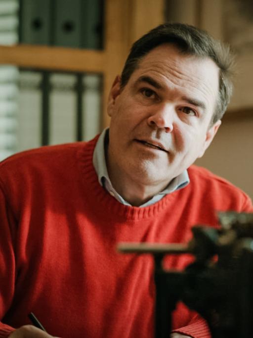 Uwe Tellkamp sitzt im roten Pullover vor einer mechanischen Schreibmaschine und scheint aufmerksam einem Gesprächspartner zuzuhören.