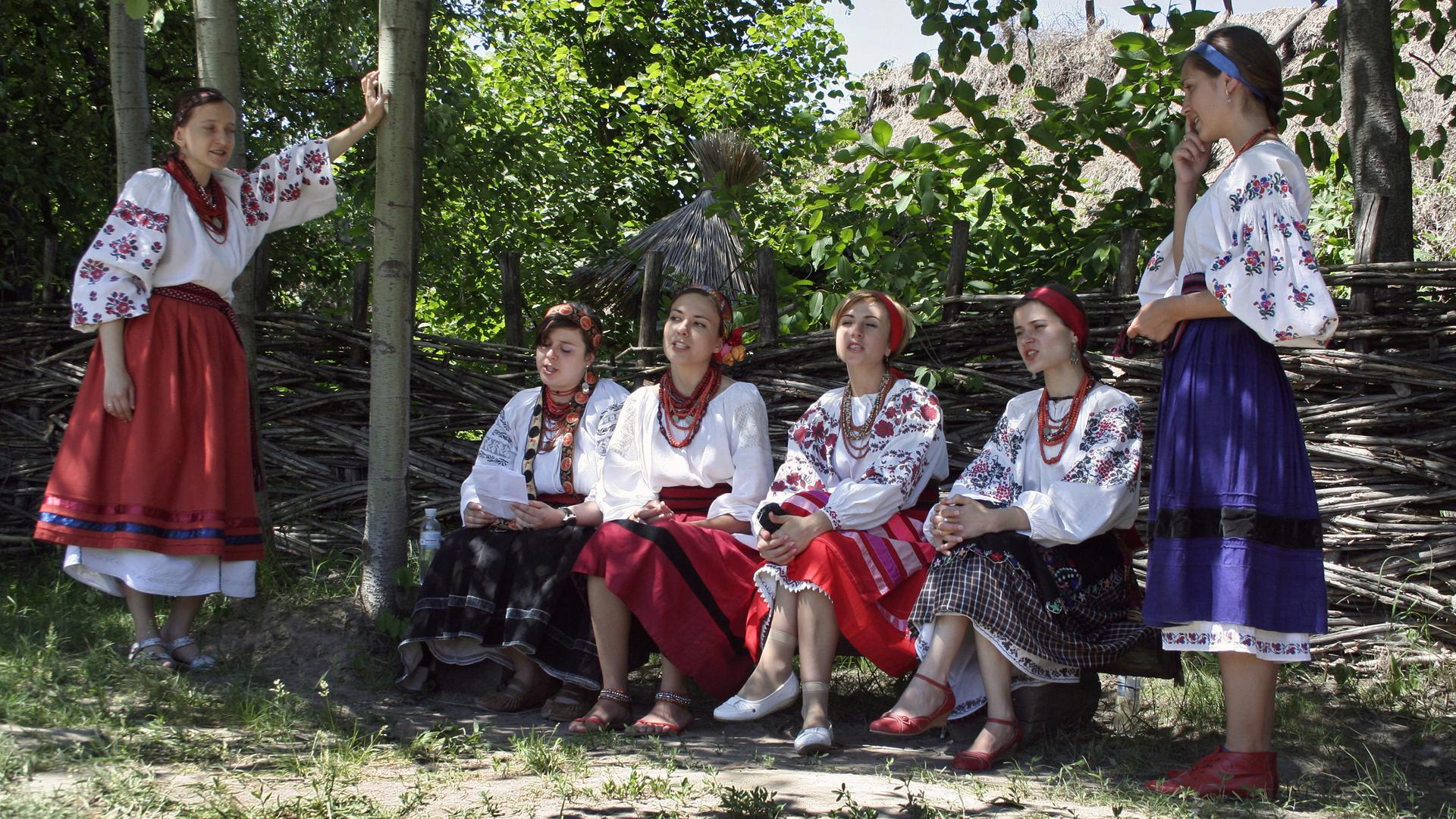 Junge ukrainische Frauen sitzen in traditioneller weißer und bunt gemusterter Kleidung auf einer Bank im Schatten und singen. Junge ukrainische Frauen sitzen in traditioneller weißer und bunt gemusterter Kleidung auf einer Bank im Schatten und singen.