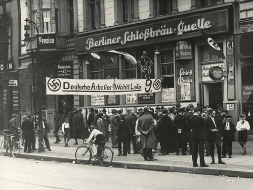 Historische Aufnahme vor einem Restaurant an dem "Berliner Schlossbräuquelle" steht. Davor ein Banner was zur Wahl der NSDAP auffordert.