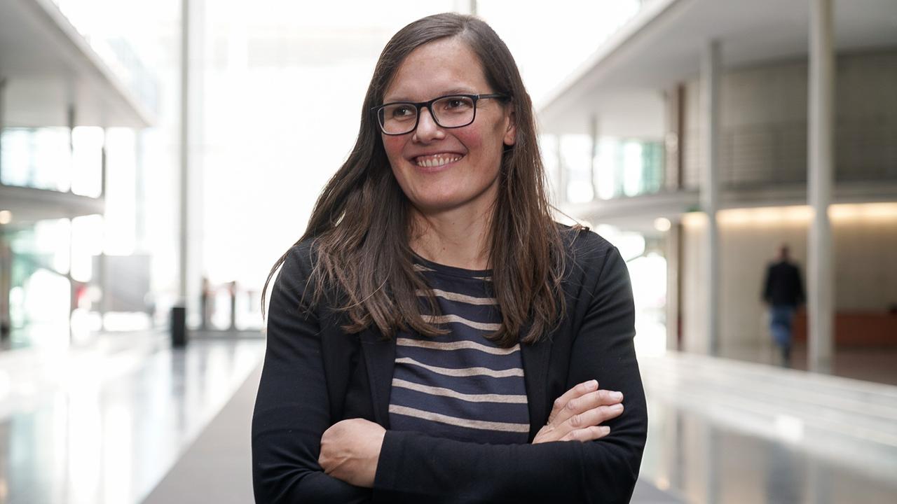 Eine Frau mit langen braunen Haaren und Brille steht in einem Bürogebäude. Eine Frau mit langen braunen Haaren und Brille steht in einem Bürogebäude.