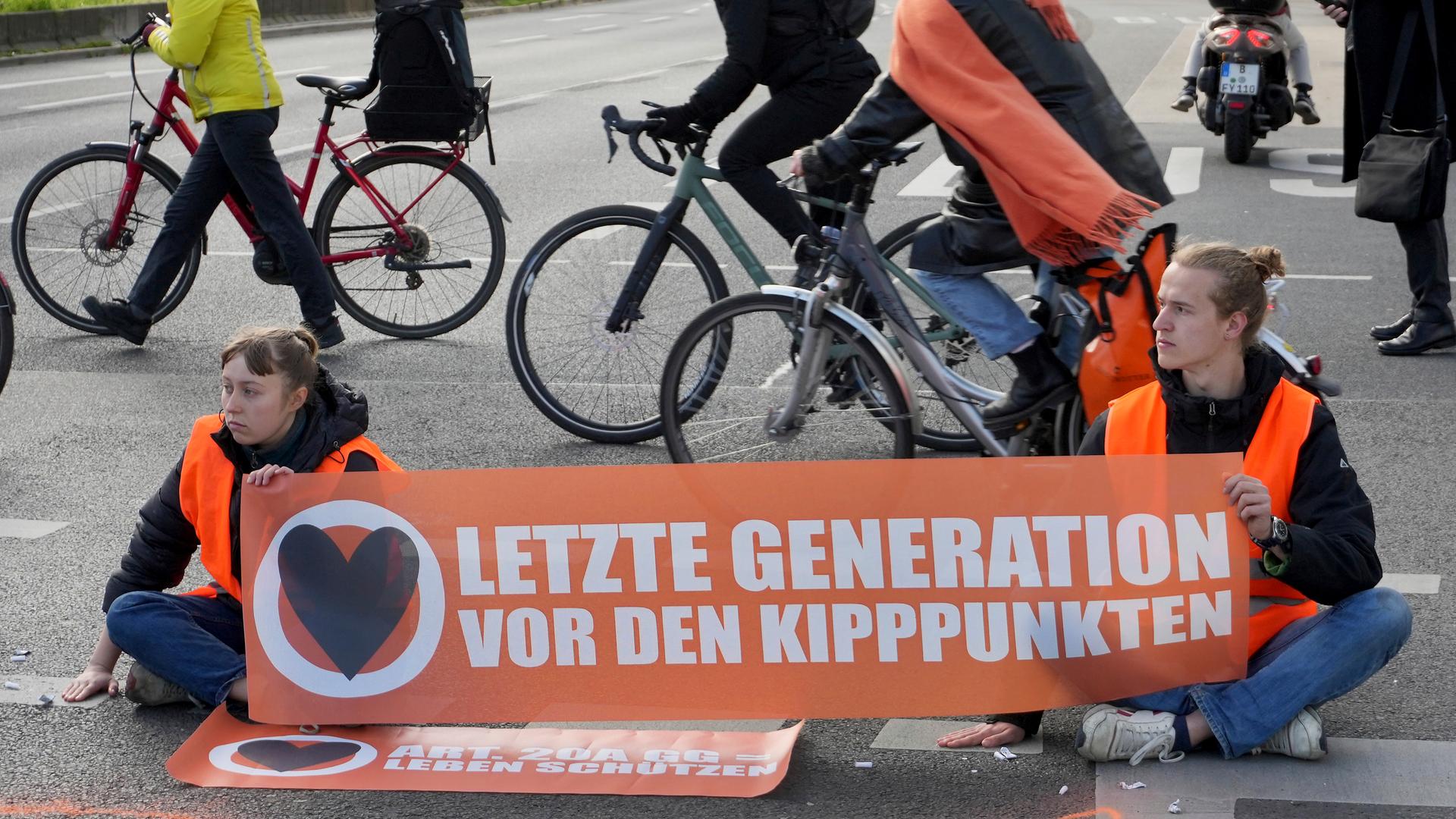 Fahrradfahrer umfahren eine Sitzblockade von zwei Klimaaktivisten der Gruppe "Letzte Generation" in Berlin. Die beiden halten ein Transparent mit der Aufschrift "Letzte Generation vor den Kipppunkten".