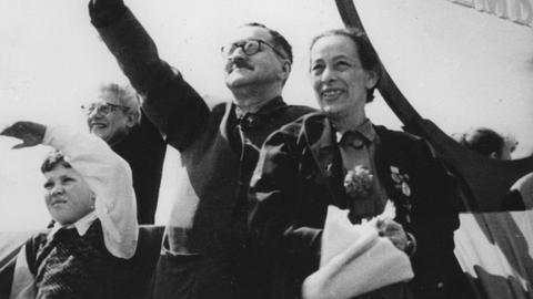 Schwarz-Weiß-Foto von Bertolt Brecht und Helene Weigel auf dem Dach des Berliner Ensembles. Er winkt mit seinem Hut, sie hält einen Blumenstrauß in der Hand.