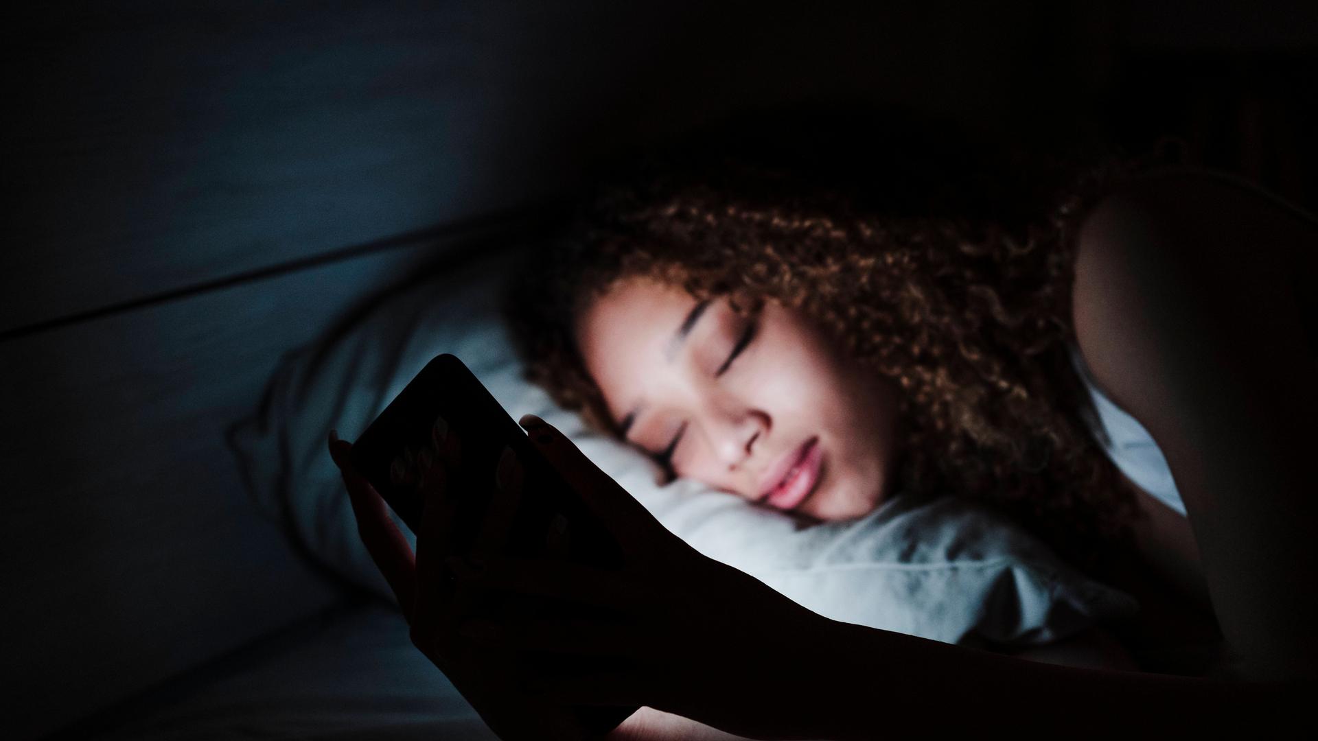 Woman holding mobile phone while sleeping on bed in dark room at home model released Symbolfoto property released EBBF03 Woman holding mobile phone while sleeping on bed in dark room at home model released Symbolfoto property released EBBF03