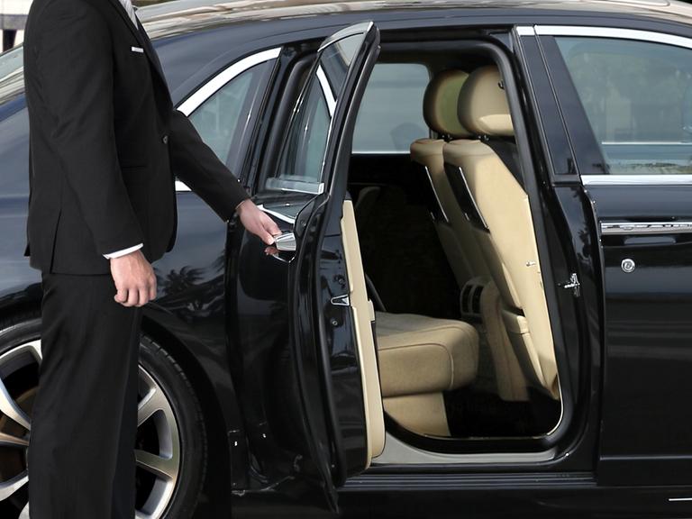 Ein Chauffeur im schwarzen Anzug öffnet die Autotür einer Luxuslimousine.