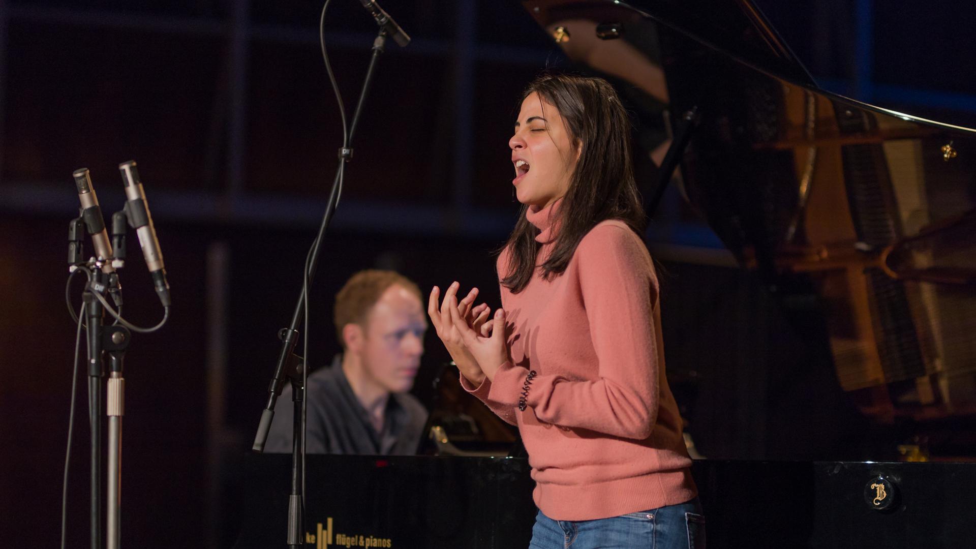 Eine junge Frau mit dunkelbraunen, schulterlangen Haaren singt ausdrucksstark. Sie formt mit den Händen eine Geste. Hinter ihr ist verschwommen der Pianist zu erkennen, der sie begleitet. Es sind die Sopranistin Fatma Said und der Pianist Joseph Middleton. 