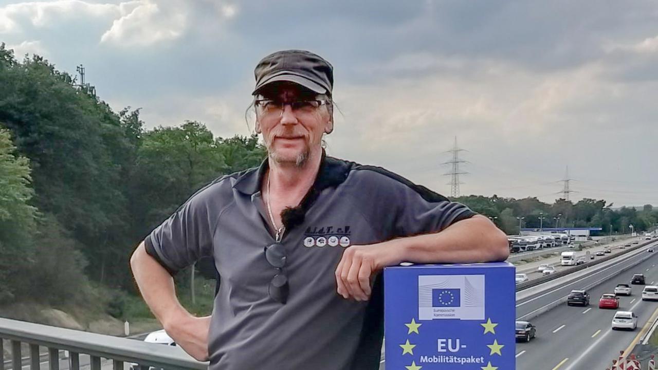 Porträt von Udo Skoppeck, der sich auf einer Autobahnbrücke ans Geländer lehnt. Auf einer blauen Box, die er hält, steht die Aufschrift "EU-Mobilitätspaket".