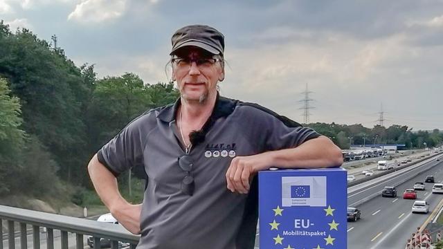 Porträt von Udo Skoppeck, der sich auf einer Autobahnbrücke ans Geländer lehnt. Auf einer blauen Box, die er hält, steht die Aufschrift "EU-Mobilitätspaket".