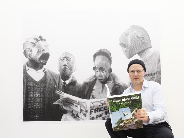 Der Künstler Jens Ullrich steht vor einem großformatigen Foto, das Menschen mit Masken zeigt