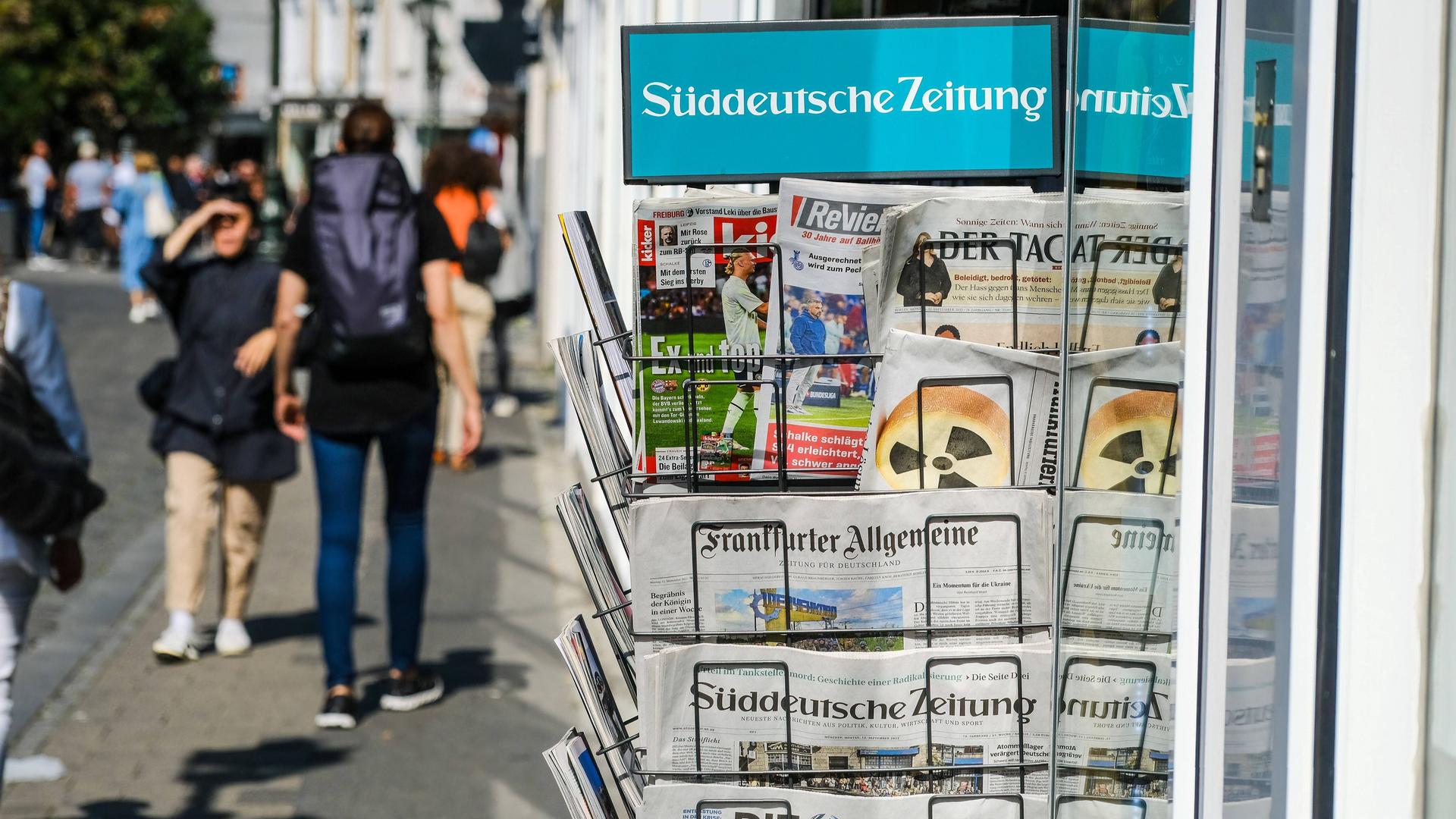 Ein Kiosk mit einem Zeitungsständer, links daneben gehen Passanten vorbei Ein Kiosk mit einem Zeitungsständer, links daneben gehen Passanten vorbei