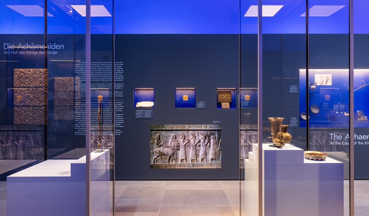 Blick in die Ausstellung „Iran. Kunst und Kultur aus fünf Jahrtausenden“, an einer Wand steht die Überschrift "Die Achämeniden", im Vordergrund sind Kunstgegenstände in Vitrinen zu sehen. Blick in die Ausstellung „Iran. Kunst und Kultur aus fünf Jahrtausenden“, an einer Wand steht die Überschrift "Die Achämeniden", im Vordergrund sind Kunstgegenstände in Vitrinen zu sehen.
