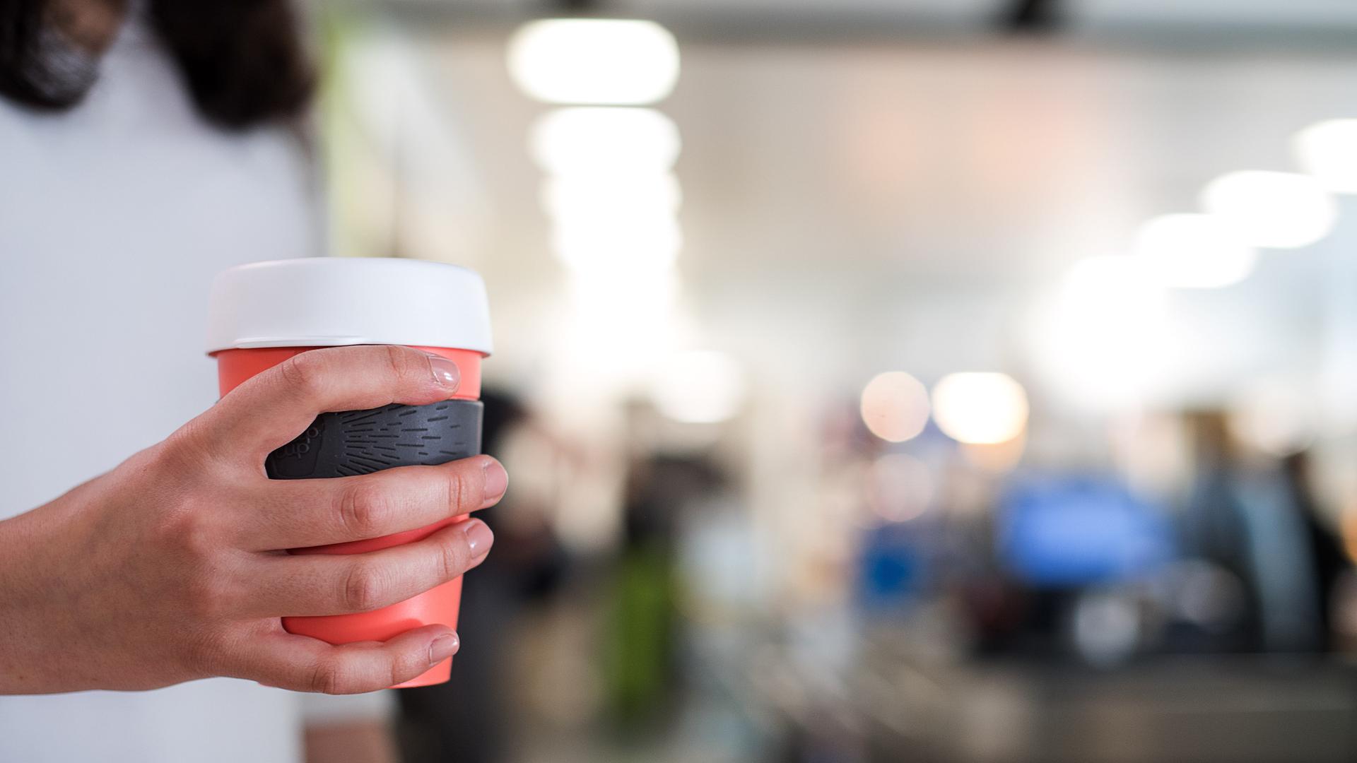 Eine Frau hält einen Mehrweg-Becher für Kaffee in der Hand. Es ist nur ihre Hand zu sehen, dahinter ein Cafe.