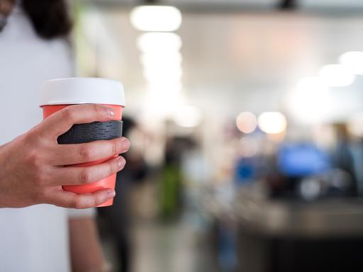 Eine Frau hält einen Mehrweg-Becher für Kaffee in der Hand. Es ist nur ihre Hand zu sehen, dahinter ein Cafe.