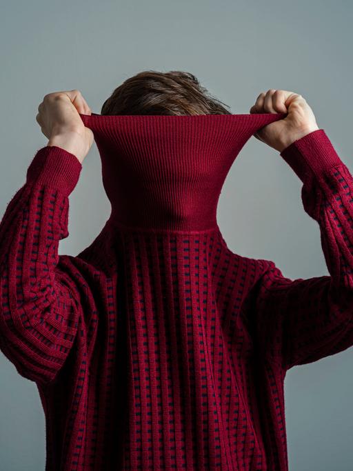 Ein Mann zieht einen weinroten Rollkragen-Pullover so über den Kopf, dass man sein Gesicht nicht mehr erkennen kann.