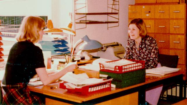 Zwei Frauen sitzen sich in einem Büro an zwei Schreibtischen gegenüber und unterhalten sich Zwei Frauen sitzen sich in einem Büro an zwei Schreibtischen gegenüber und unterhalten sich