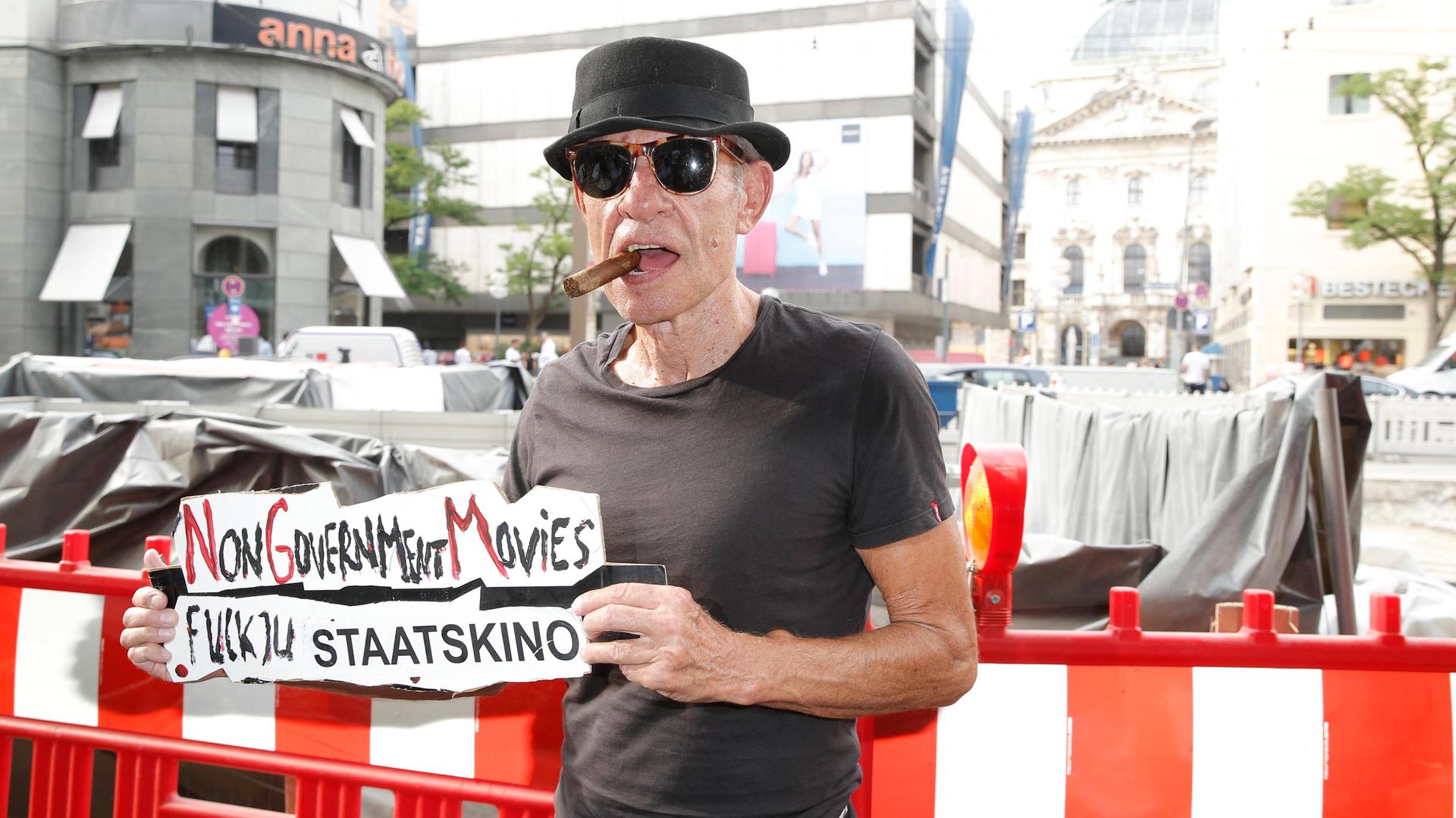 Klaus Lemke steht mit einer Zigarre im Mund und mit Sonnenbrille auf der Straße und hält ein Schild hoch, auf dem steht "Fuck You Staatskino"