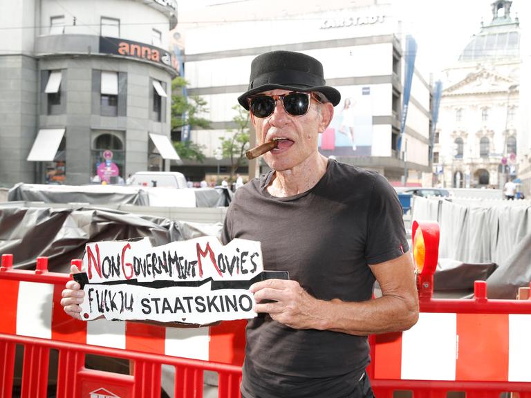 Klaus Lemke steht mit einer Zigarre im Mund und mit Sonnenbrille auf der Straße und hält ein Schild hoch, auf dem steht "Fuck You Staatskino"