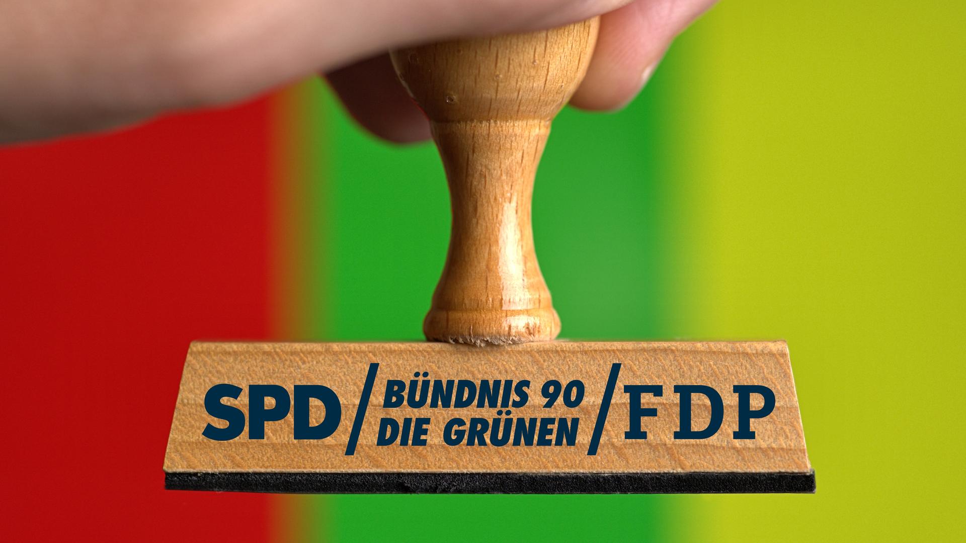 Eine Hand hält einen Holzstempel, auf dem die Parteilogos von SPD, Bündnis 90/Die Grünen und FDP stehen. Der Hintergrund ist rot-grün-gelb gestreift.