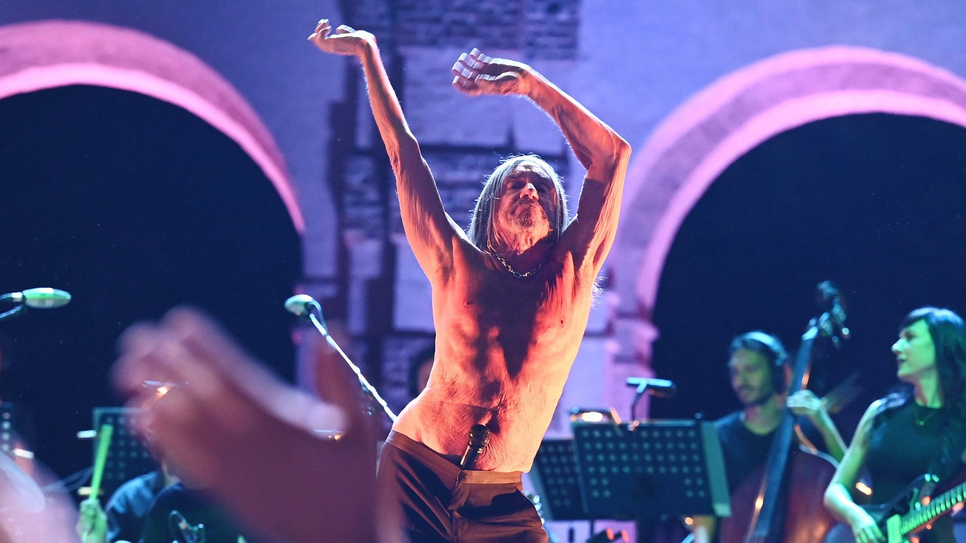 Der 75-jährige Sänger Iggy Pop tanzt mit freiem Oberkörper während eines Konzerts auf der Bühne des Palazzo Te in Mantua. In der Hüfte knickt er seitlich ab, die Hände hat er hoch in der Luft, das Mikrofon steckt im Hosenbund. Der 75-jährige Sänger Iggy Pop tanzt mit freiem Oberkörper während eines Konzerts auf der Bühne des Palazzo Te in Mantua. In der Hüfte knickt er seitlich ab, die Hände hat er hoch in der Luft, das Mikrofon steckt im Hosenbund.