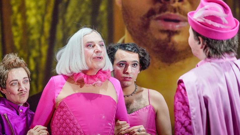Drei Darsteller in rosa und lila Kostümen stehen in Richtung Kamera, einer mit rosa Hut wendet uns den Rücken zu. Im Hintergrund ist ein bärtiges Gesicht auf eine Leinwand projiziert. Drei Darsteller in rosa und lila Kostümen stehen in Richtung Kamera, einer mit rosa Hut wendet uns den Rücken zu. Im Hintergrund ist ein bärtiges Gesicht auf eine Leinwand projiziert.