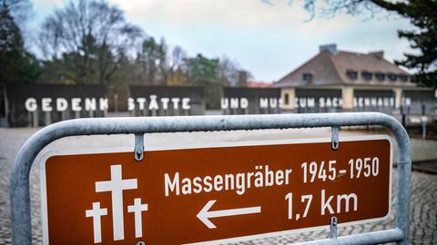 Die KZ-Gedänkstätte Sachsenhausen. Auf einem Schild im Vordergrund steht "Massengräber 1945-1950 in 1,7 km". Im Hintergrund sieht man den Eingang zur Gedenkstätte mit dem Schriftzug "Gedenkstätte und Museum Sachsenhausen".