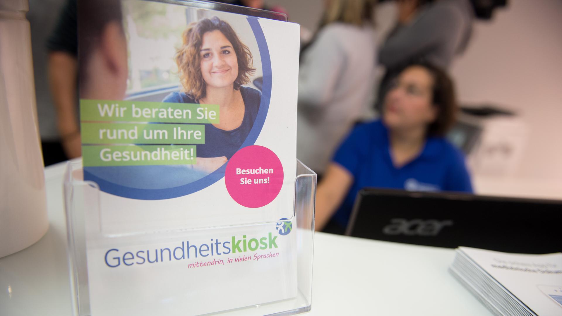 Das Foto zeigt einen Aufsteller mit bunten Broschüren, auf denen eine freundlich lächelnde Frau abgebildet ist. Es sind Infoblätter des Gesundheitskiosk in Hamburg-Billstedt. Im Hintergrund ist leicht unscharf eine Pflegekraft im blauen Kittel zu sehen. 