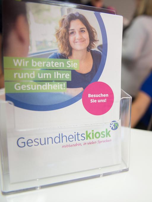 Das Foto zeigt einen Aufsteller mit bunten Broschüren, auf denen eine freundlich lächelnde Frau abgebildet ist. Es sind Infoblätter des Gesundheitskiosk in Hamburg-Billstedt. Im Hintergrund ist leicht unscharf eine Pflegekraft im blauen Kittel zu sehen. 