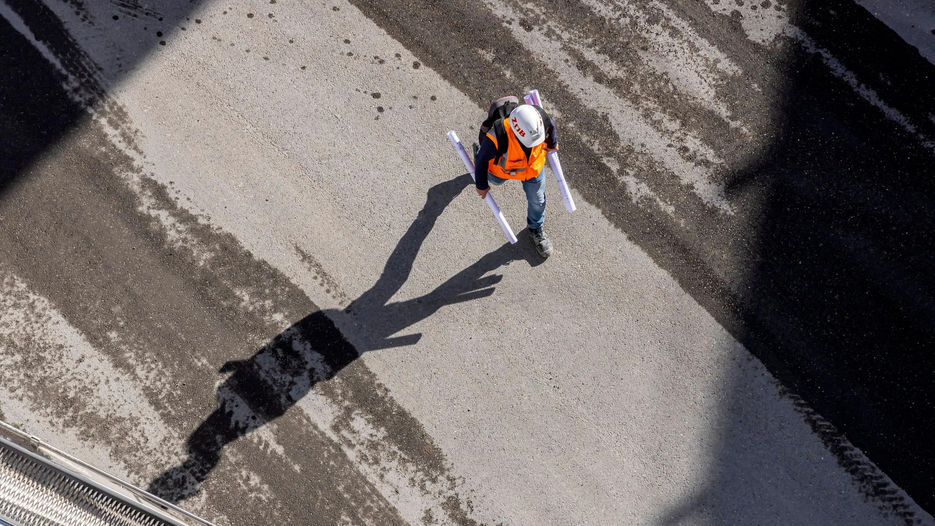 Eine Baustelle in Stuttgart: Ein Bauarbeiter mit Helm und Schutzweste trägt zwei Papierrollen. Eine Baustelle in Stuttgart: Ein Bauarbeiter mit Helm und Schutzweste trägt zwei Papierrollen.