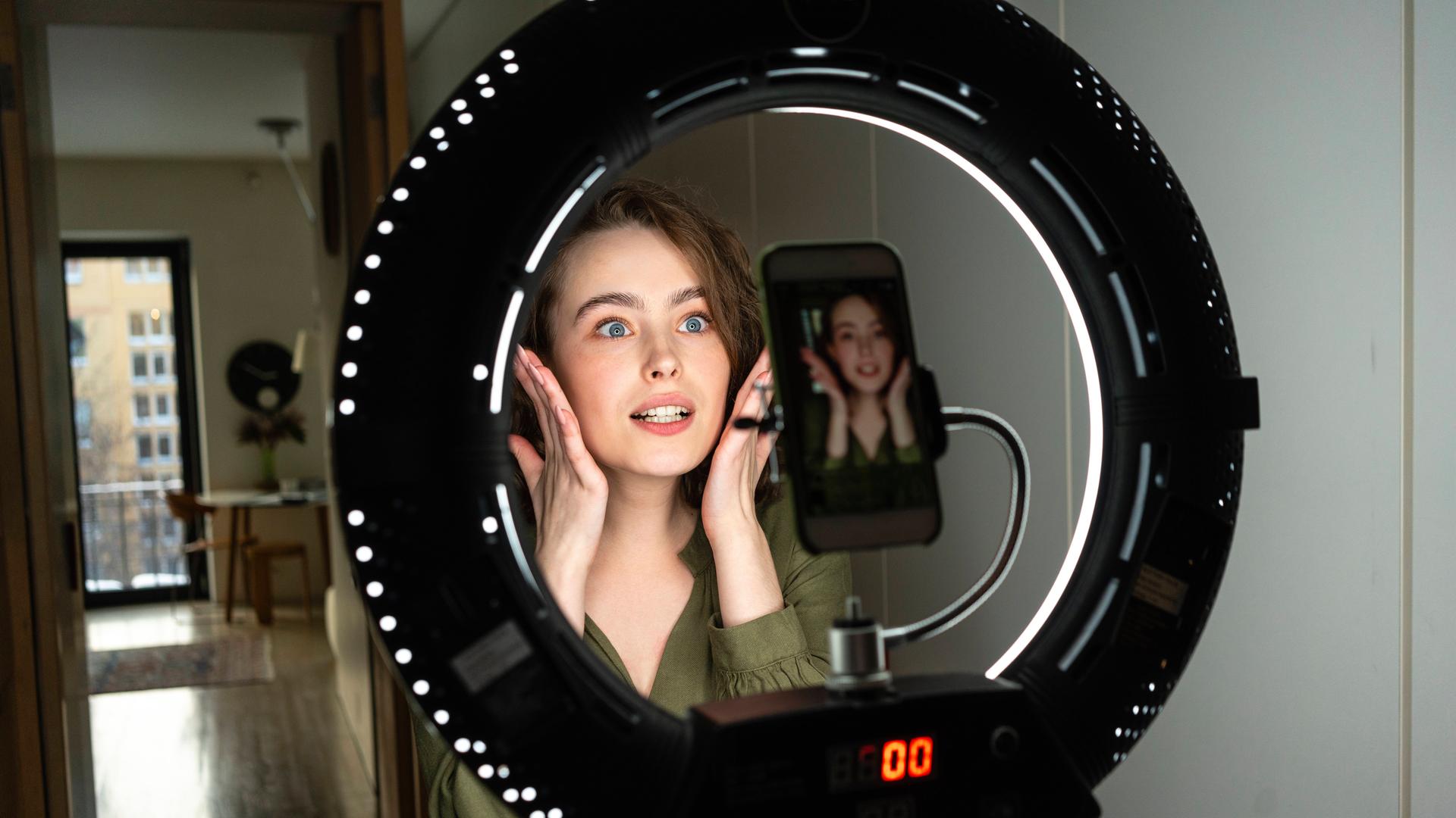 Eine junge Frau posiert vor ihrer Smartphonekamera, die in der Mitte eines Ringlichts montiert ist.