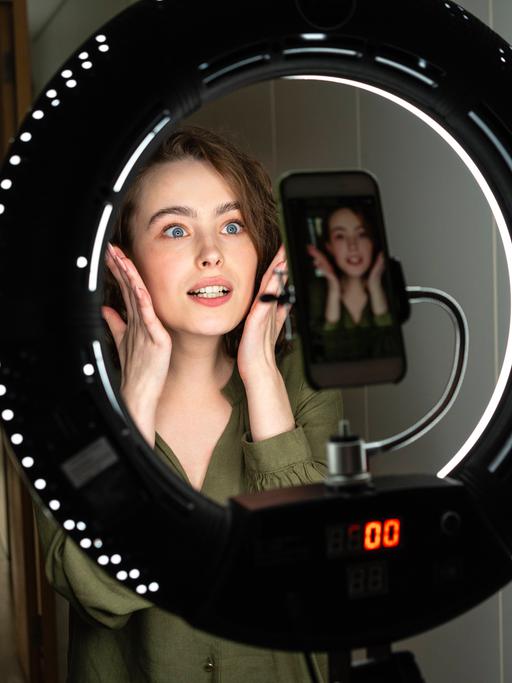 Eine junge Frau posiert vor ihrer Smartphonekamera, die in der Mitte eines Ringlichts montiert ist.