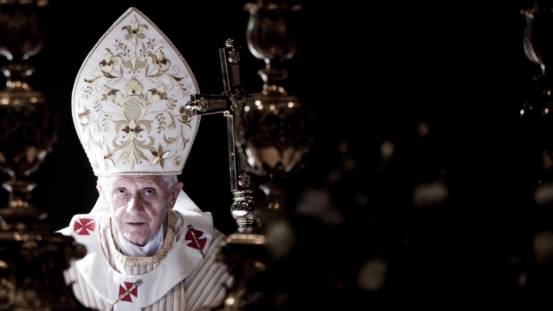 Papst Benedikt trägt eine weiße, oben spitz zulaufende Kopfbedeckung mit goldenen Ornamenten und schaut nach oben.