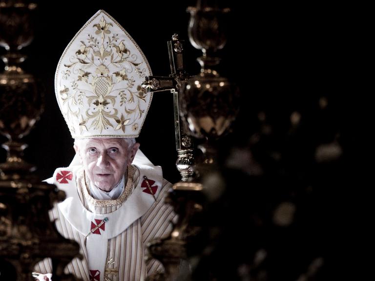 Papst Benedikt trägt eine weiße, oben spitz zulaufende Kopfbedeckung mit goldenen Ornamenten und schaut nach oben.