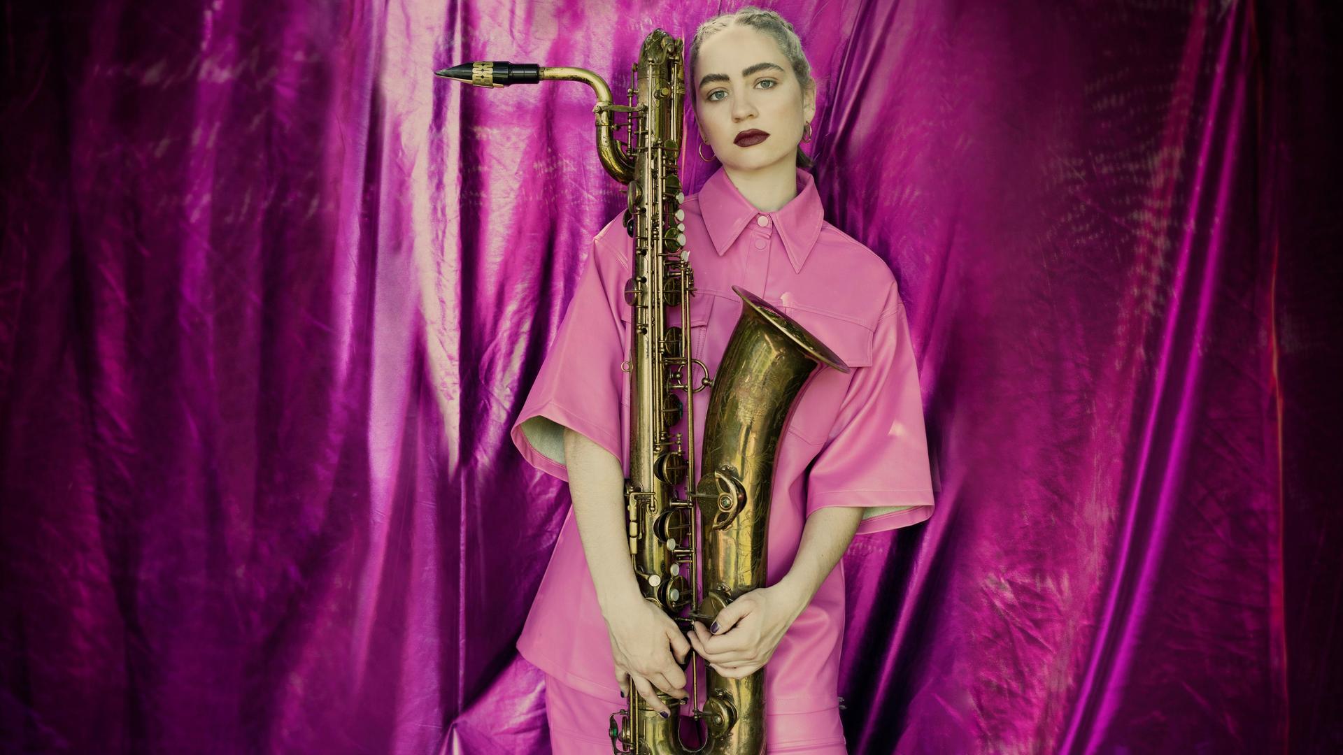 Eine junge Frau steht komplett in rosa gekleidet mit einem Saxofon in der Hand vor einem pinken Vorhang.