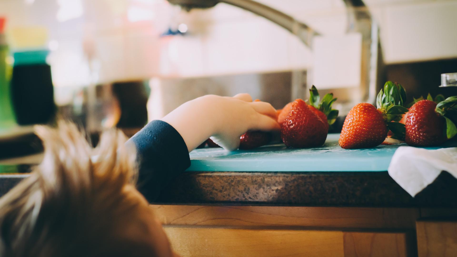 Ein Kind greift von unten nach Erbeeren, die auf einer Küchenarbeitsplatte liegen. Ein Kind greift von unten nach Erbeeren, die auf einer Küchenarbeitsplatte liegen.