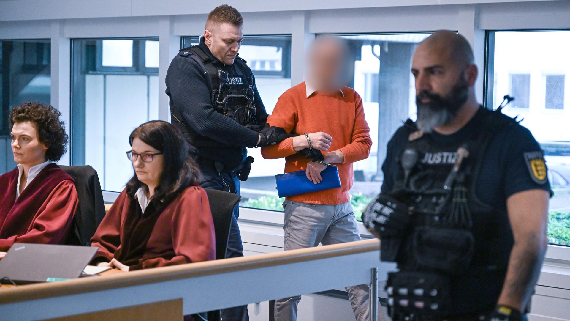Ein Mann mit verpixeltem Gesicht wird in Handschellen von einem uniformierten Justizbeamten in einen Gerichtssal geführt. Rechts steht ein weiterer Beamter. Ein Mann mit verpixeltem Gesicht wird in Handschellen von einem uniformierten Justizbeamten in einen Gerichtssal geführt. Rechts steht ein weiterer Beamter.
