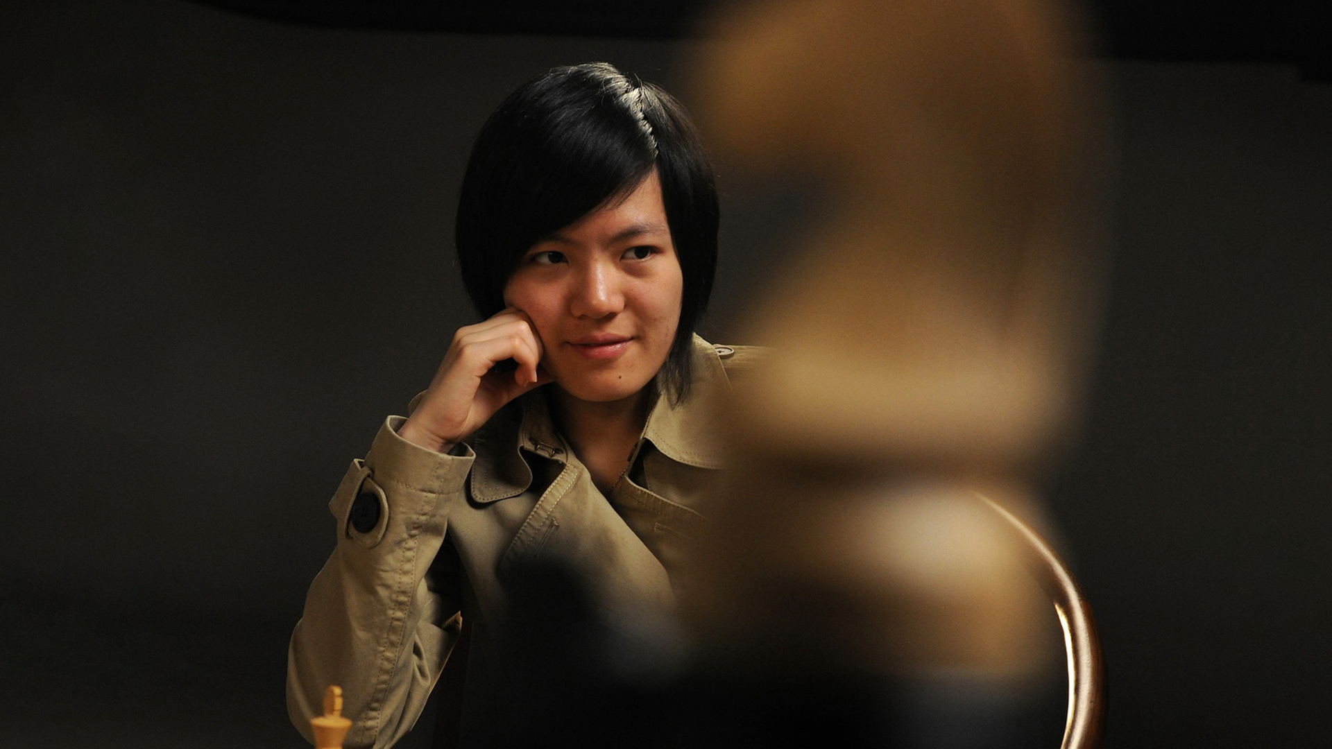 Hou Yifan sitzt vor einem schwarzen Hintergrund und trägt Trenchcoat. Sie blickt herausfordernd auf einen Schach-Springer, der sich unscharf im Vordergrundgrund befindet und perspektivisch das Bild bestimmt Hou Yifan sitzt vor einem schwarzen Hintergrund und trägt Trenchcoat. Sie blickt herausfordernd auf einen Schach-Springer, der sich unscharf im Vordergrundgrund befindet und perspektivisch das Bild bestimmt