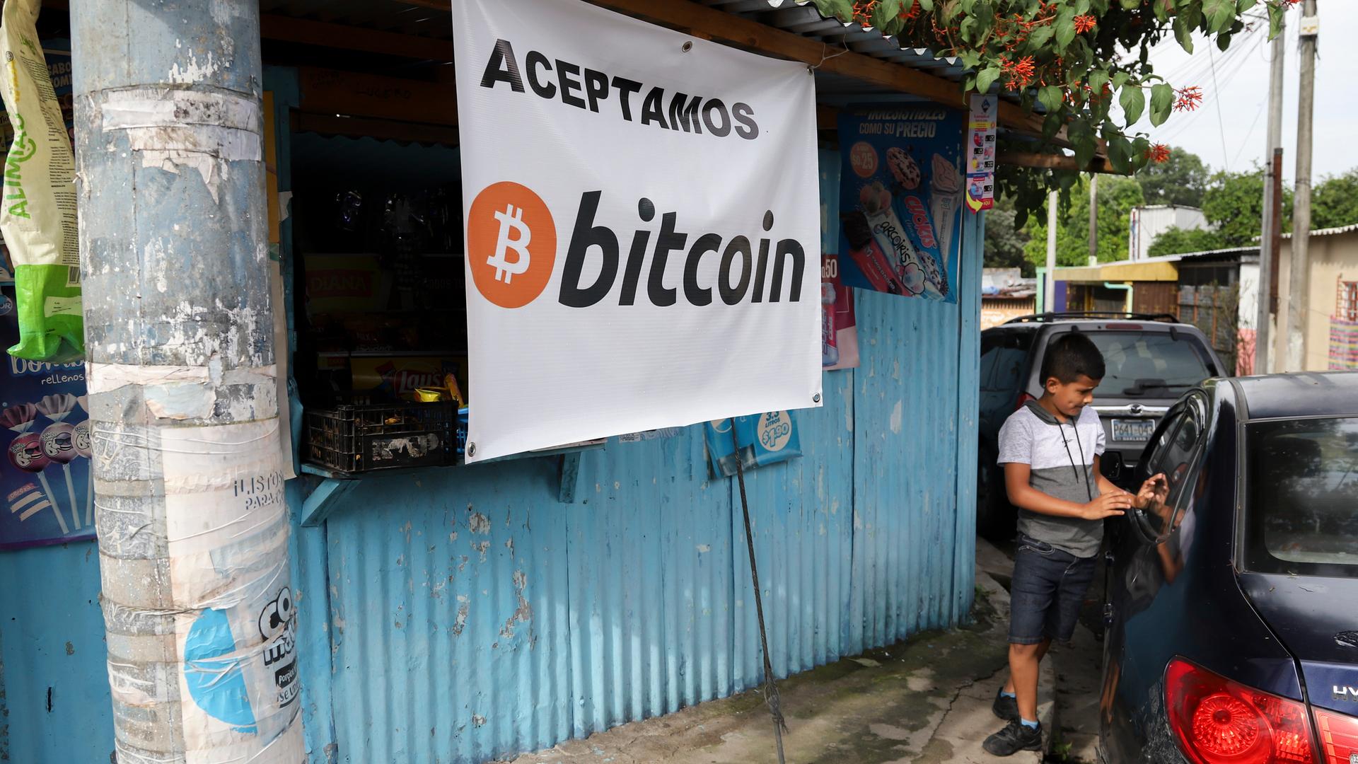 Ein Jugendlicher steht neben einem Auto, während er vor einem kleinen Geschäft wartet, das Bitcoin akzeptiert. Ein Jugendlicher steht neben einem Auto, während er vor einem kleinen Geschäft wartet, das Bitcoin akzeptiert.