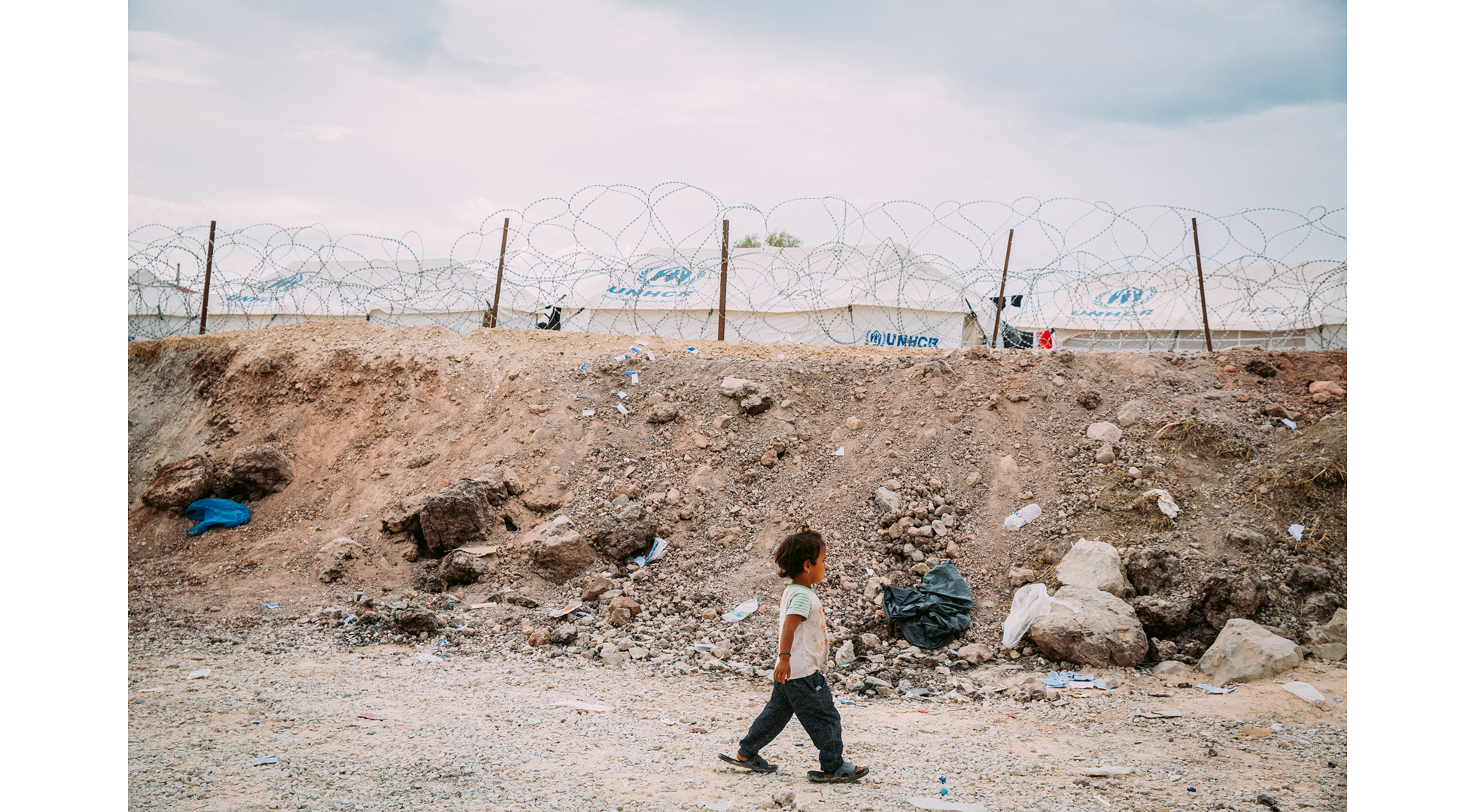 Ein Junge in zu großen Schuhen geht an einem Stacheldrahtzaun entlang, dahinter stehen Zelte mit der Aufschrift UNHCR 