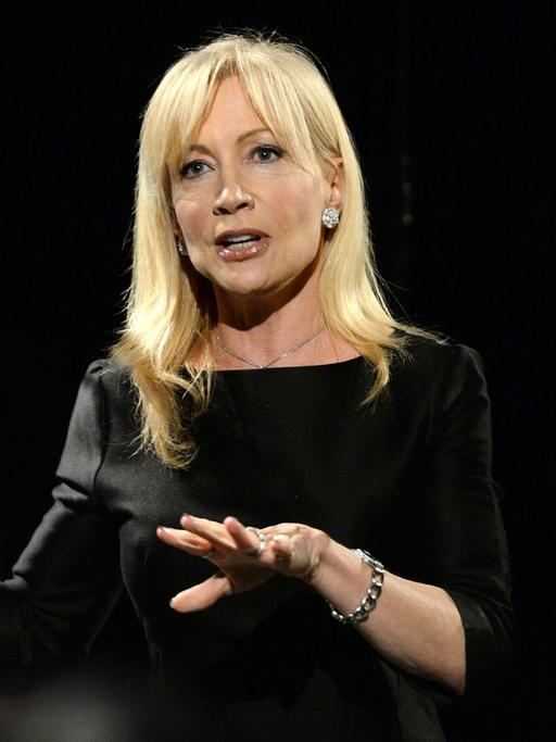 Eine Frau mit offenen blonden Haaren steht in einem edlen, schwarzen Kleid auf der Bühne und hebt ihre Hände.