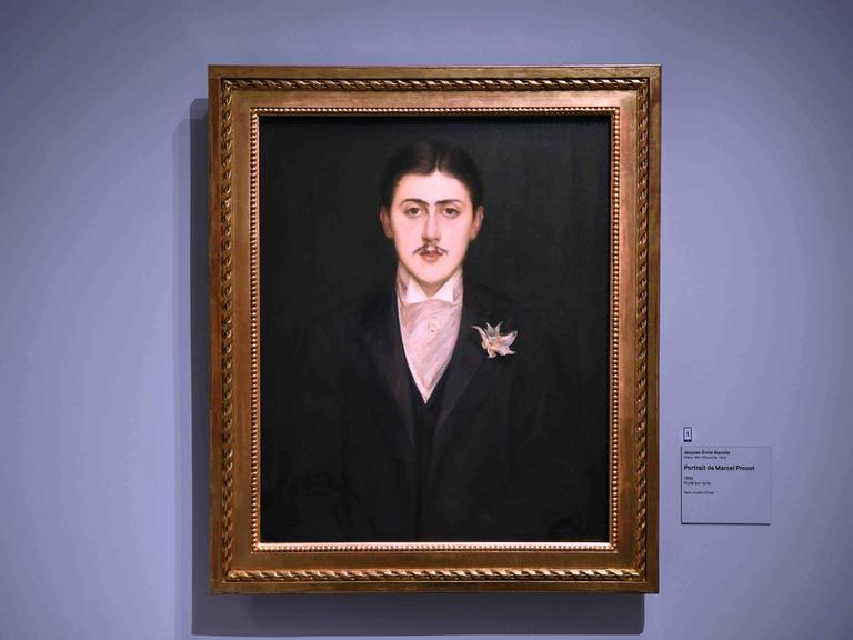 Ein gerahmtes Portraitbild des französischen Schriftstellers Marcel Proust hängt in einem Rahmen an einer violetten Wand. Daneben hängt eine Beschriftung des Bildes.