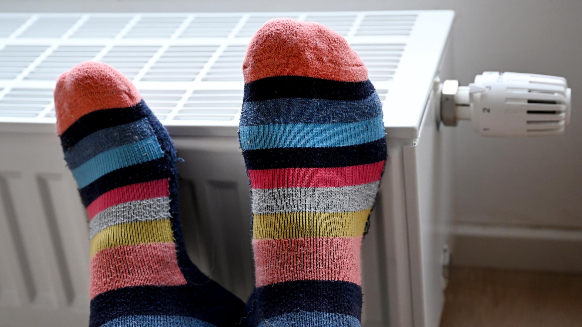 Füße in geringelten Socken liegen zum Wärmen auf einem Heizkörper in einer Wohnung. Füße in geringelten Socken liegen zum Wärmen auf einem Heizkörper in einer Wohnung.