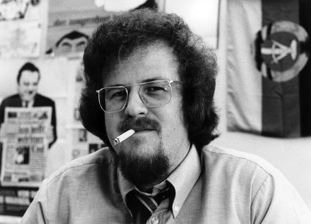 Jörg Mettke. Ein Mann mit lockigen Haaren und Brille sitzt in einem Büro und hat eine brennende Zigarette im Mund. Im Hintergrund hängen an der Wand eine DDR-Flagge und ein Bild von Franz Josef Strauß. Jörg Mettke. Ein Mann mit lockigen Haaren und Brille sitzt in einem Büro und hat eine brennende Zigarette im Mund. Im Hintergrund hängen an der Wand eine DDR-Flagge und ein Bild von Franz Josef Strauß.
