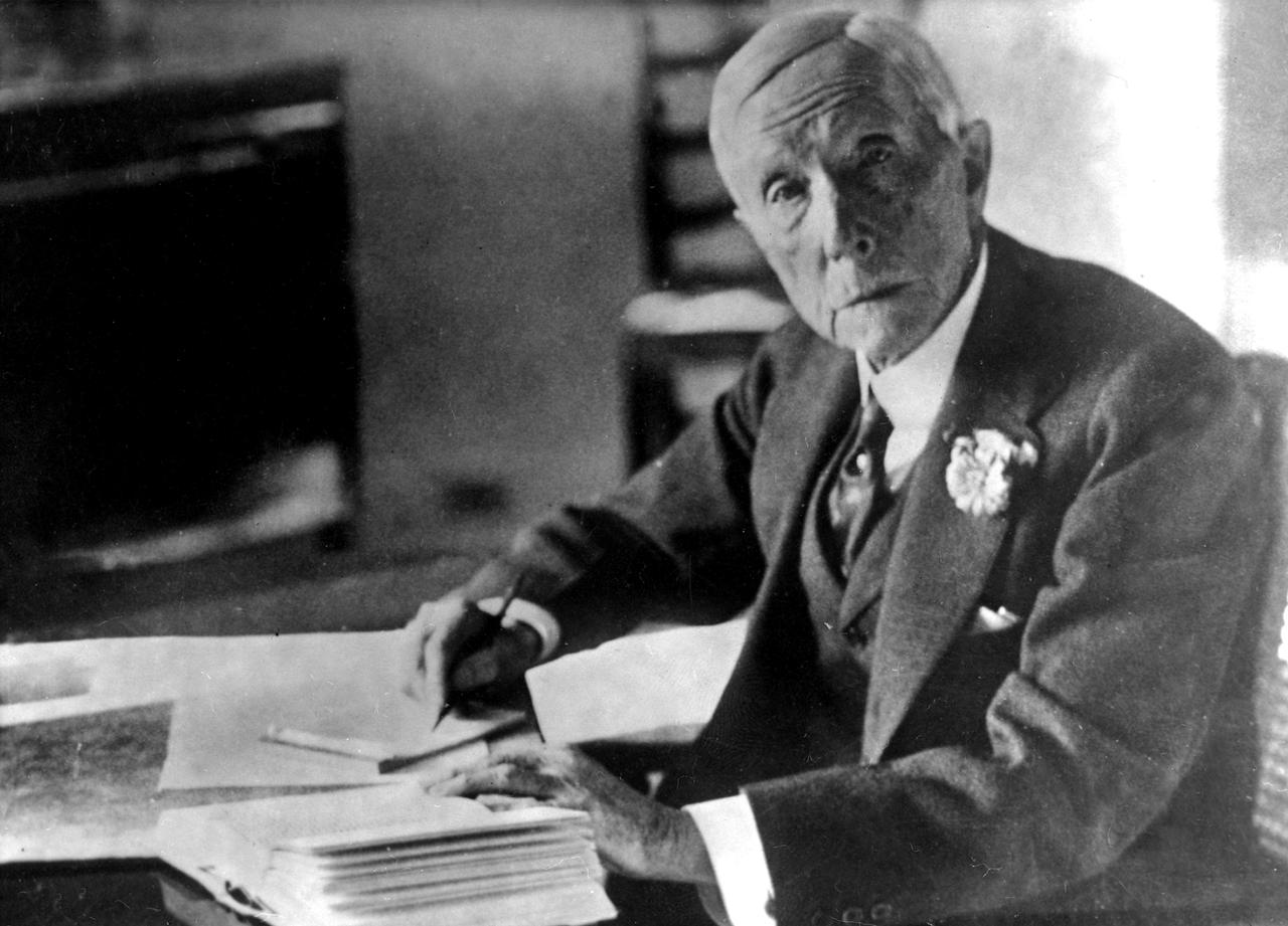 Der amerikanische Unternehmer und Ölmagnat John D. Rockefeller sitzt an einem Schreibtisch und guckt in die Kamera. In der rechten Hand hält er einen Stift.