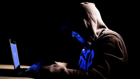 Ein junger Mann mit schwarzem Kapuzenpullover sitzt im Dunkeln vor dem Bildschirm eines Laptops, der blaues Licht auf sein halb verdecktes Gesicht wirft.