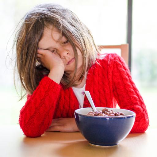 Ein müdes Kind sitzt mit geschlossenen Augen und aufgestütztem Kopf vor einer Schüssel Frühstücksflocken