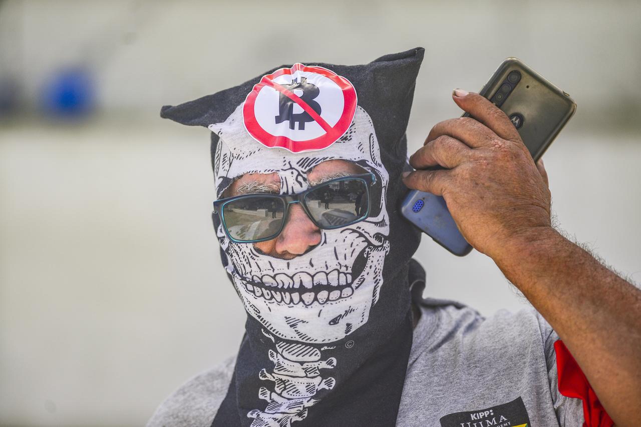 Ein Demonstrant telefoniert während des Protestmarsches, während er eine Maske mit Anti-Bitcoin-Bildern trägt. Ein Demonstrant telefoniert während des Protestmarsches, während er eine Maske mit Anti-Bitcoin-Bildern trägt.