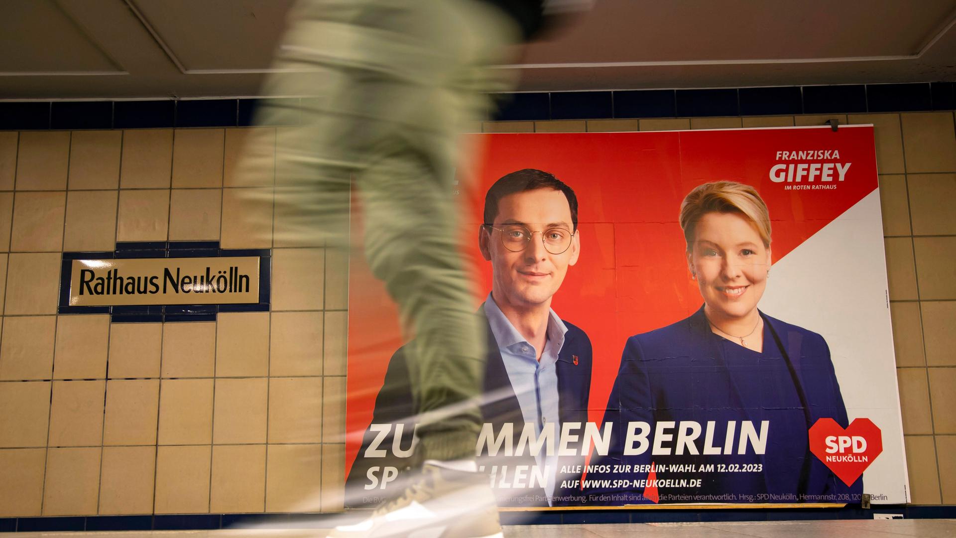 Ein Wahlplakat von Franziska Giffey und Martin Hikel hängt in der U-Bahn-Station Neukölln. Im Vordergrund sieht man mit Bewegungsunschärfe ein Bein durchs Bild laufen.